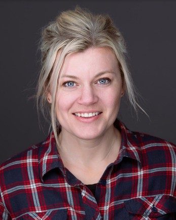 A photo of the artist Andi Burnum. She's a blonde woman wearing a plaid shirt, smiling at the camera and standing against a dark grey background.